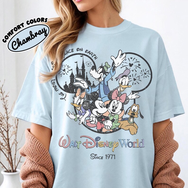 May include: Light blue t-shirt featuring a graphic of Disney characters including Mickey, Minnie, Donald, Daisy, Goofy, and Pluto. The design includes the text "Walt Disney World" and "Since 1971". The shirt is labeled "Comfort Colors Chambray".