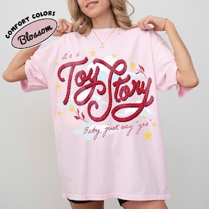 May include: A light pink t-shirt featuring the text 'it's a Toy Story' in a red rope-style font, accompanied by yellow stars, light blue clouds, and small floral accents. Below the main text, the phrase 'Baby, just say "yes"' is printed in red.