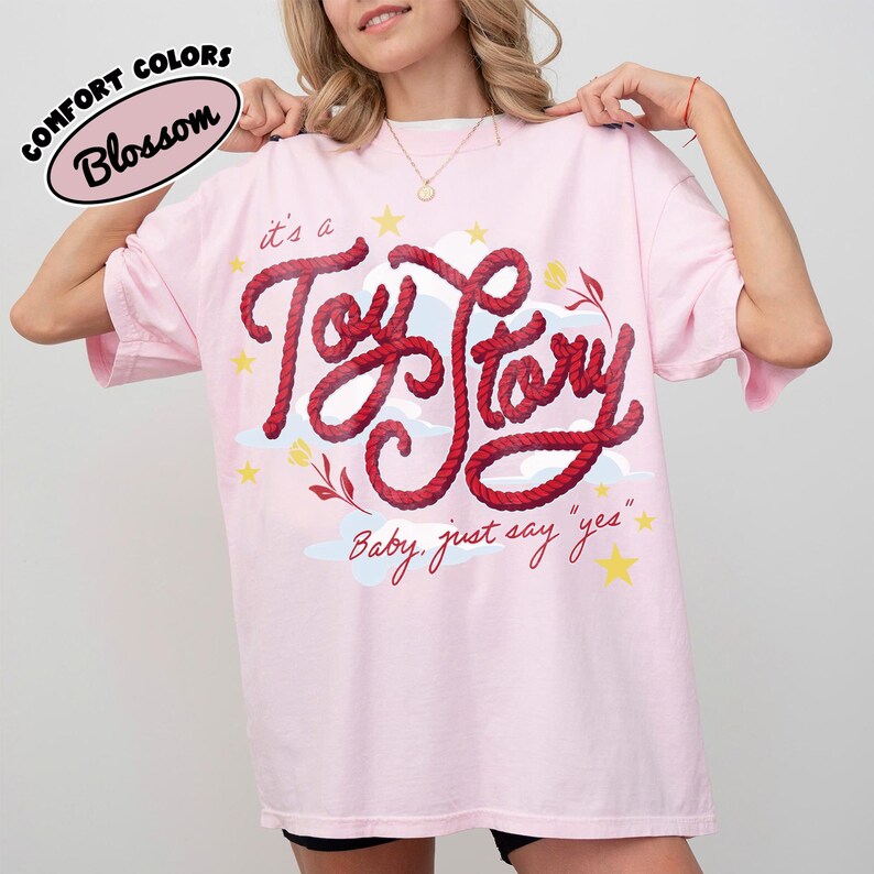 May include: A light pink t-shirt featuring the text 'it's a Toy Story' in a red rope-style font, accompanied by yellow stars, light blue clouds, and small floral accents. Below the main text, the phrase 'Baby, just say "yes"' is printed in red.