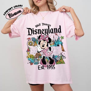 Op de afbeelding: Een lichtroze t-shirt met een afbeelding van Minnie Mouse in het midden, omringd door diverse Disneyland-attracties. De tekst luidt Walt Disney's Disneyland Est. 1955. Een klein ovaal logo linksboven vermeldt Comfort Colors Blossom.