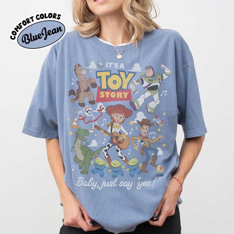 May include: A blue short-sleeve t-shirt featuring a graphic print of Toy Story characters including Woody, Jessie, Buzz Lightyear, Rex, Bullseye, Forky, and the Aliens. The text reads IT'S A TOY STORY and Baby, just say 'yes!' with a logo at the top.