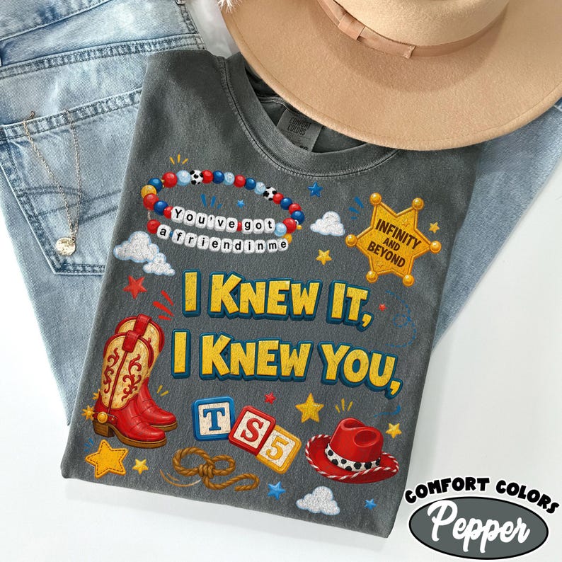 May include: A charcoal gray t-shirt featuring a graphic print with red cowboy boots, a red cowboy hat, a beaded bracelet with the text You've got a friend in me, a sheriff badge with Infinity and Beyond, and blocks with TS5, plus yellow stars.