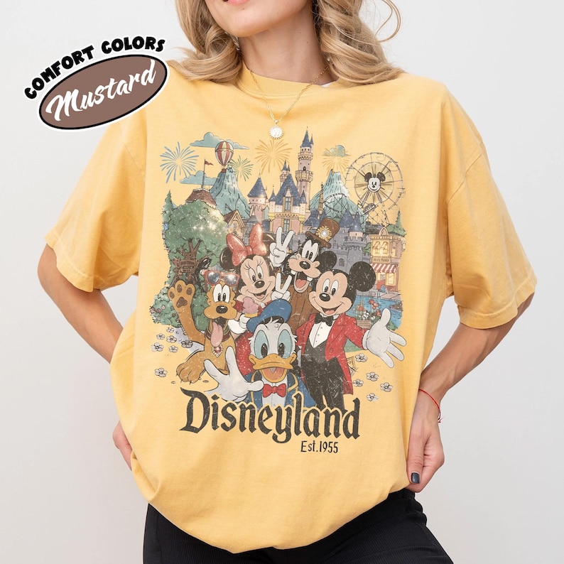 May include: Mustard-colored t-shirt featuring a Disneyland graphic with Mickey Mouse, Minnie Mouse, Donald Duck, Goofy, and other characters. The graphic includes a castle, fireworks, and the text "Disneyland Est. 1955". The shirt has short sleeves and a crew neck.
