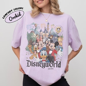 May include: A light purple t-shirt with a graphic of Disney characters in front of a castle and fireworks. The word "Disneyworld" is printed in a vintage font, with "Est.1971" below. The shirt has the text "Comfort Colors" and "Orchid" printed on it.