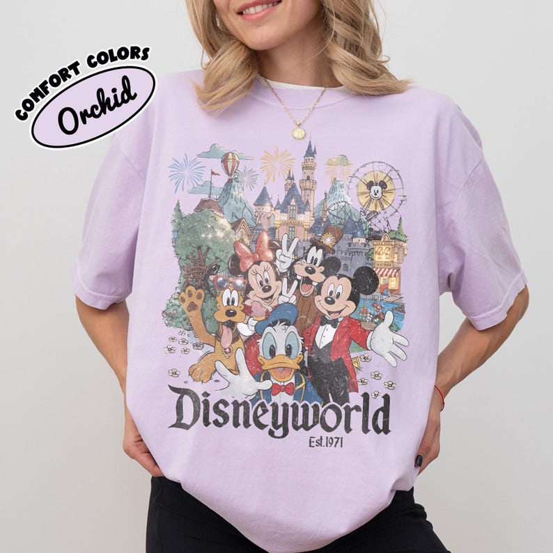 May include: A light purple t-shirt with a graphic of Disney characters in front of a castle and fireworks. The word "Disneyworld" is printed in a vintage font, with "Est.1971" below. The shirt has the text "Comfort Colors" and "Orchid" printed on it.