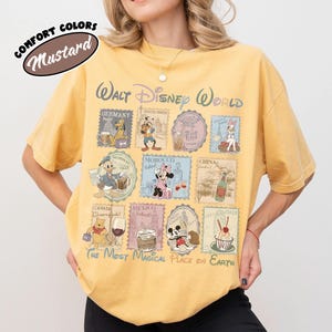 May include: Mustard-colored t-shirt featuring a Walt Disney World design. The shirt has a stamp-like graphic with Disney characters and country names. Text includes "The Most Magical Place on Earth."