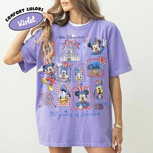 May include: Violet Comfort Colors t-shirt featuring a colorful Disney World design. The design includes images of Mickey Mouse, Minnie Mouse, Donald Duck, and Goofy, along with the text "250 years of freedom" and "2026". The shirt has a crew neck and short sleeves.