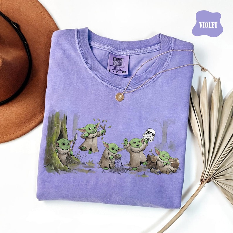 May include: A violet short-sleeve t-shirt featuring a graphic print of five small green creatures in tan robes. One creature holds a spear, one a slingshot, one a net, one a white helmet, and one sits on a log. The shirt has a Comfort Colors label.
