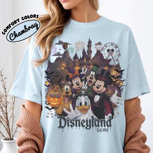 May include: A light blue t-shirt featuring a graphic of Disney characters in Halloween costumes, including Mickey Mouse, Minnie Mouse, Donald Duck, Goofy, and Pluto. The design includes a castle, a ferris wheel, fireworks, and the text Disneyland Est. 1955.