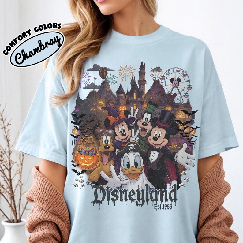 May include: A light blue t-shirt featuring a graphic of Disney characters in Halloween costumes, including Mickey Mouse, Minnie Mouse, Donald Duck, Goofy, and Pluto. The design includes a castle, a ferris wheel, fireworks, and the text Disneyland Est. 1955.