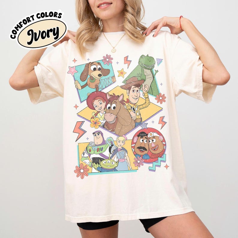 Puede incluir: Camiseta de color marfil con un gr&aacute;fico colorido de personajes animados. El dise&ntilde;o incluye un perro, un dinosaurio, un vaquero, un caballo y otros personajes. La camiseta tiene las palabras "Comfort Colors" e "Ivory" impresas.