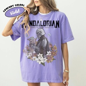 May include: Violet Comfort Colors t-shirt featuring a graphic of The Mandalorian and Grogu surrounded by flowers and butterflies. The text "MANDALORIAN" and "THIS IS THE WAY" are printed above the graphic.