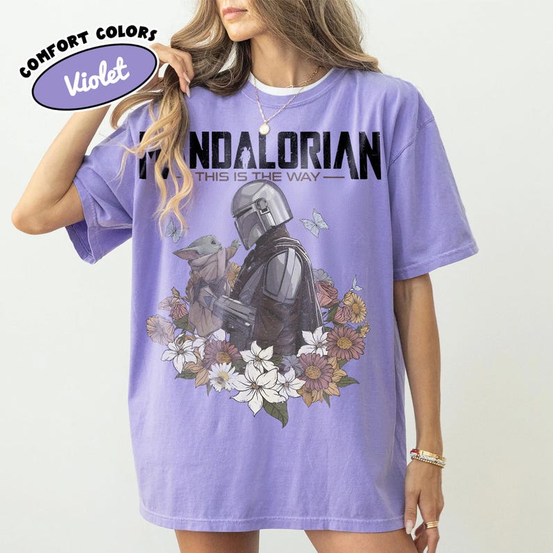 May include: Violet Comfort Colors t-shirt featuring a graphic of The Mandalorian and Grogu surrounded by flowers and butterflies. The text "MANDALORIAN" and "THIS IS THE WAY" are printed above the graphic.