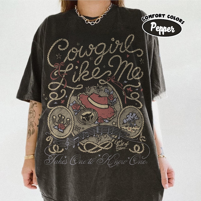 May include: A charcoal gray short-sleeve t-shirt featuring a graphic print with the text Cowgirl Like Me, Eyes full of stars, and Takes One to Know One. The design includes a red hat, a horseshoe, a bull skull, and rope motifs.