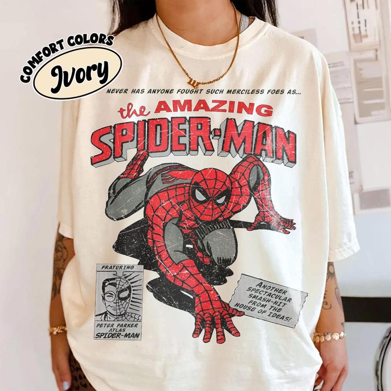 May include: An ivory t-shirt featuring a vintage-style graphic of Spider-Man in a crouching pose. The text reads: NEVER HAS ANYONE FOUGHT SUCH MERCILESS FOES AS... THE AMAZING SPIDER-MAN, FRATURING PETER PARKER ATLAS SPIDER-MAN, and ANOTHER SPECTACULAR SMASH-HIT FROM THE HOUSE OF IDEAS!