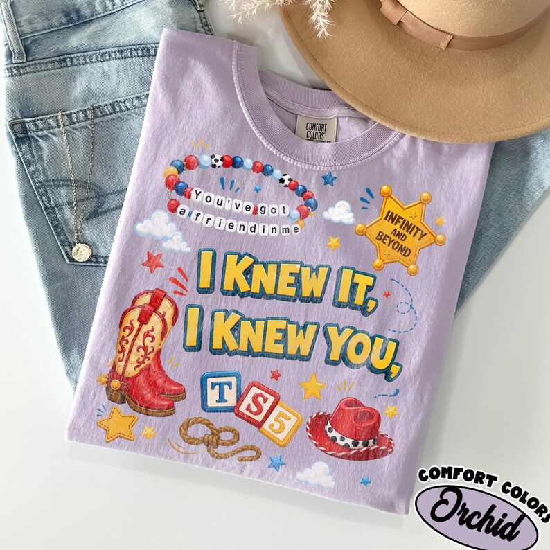 May include: A light purple t-shirt featuring a graphic print with a beaded bracelet, a yellow sheriff star badge with the text INFINITY AND BEYOND, red cowboy boots, alphabet blocks spelling TS5, a lasso, and a red cowboy hat with the text I KNEW IT, I KNEW YOU.