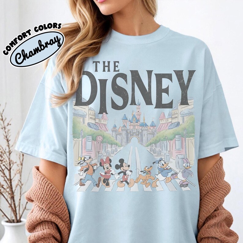 Może przedstawiać: Jasnoniebieska koszulka z szarym napisem "THE DISNEY". Koszulka przedstawia kolorową ilustrację postaci Disneya przechodzących przez ulicę z zamkiem w tle. Słowa "COMFORT COLORS Chambray" znajdują się w okręgu po lewej stronie u g&oacute;ry.