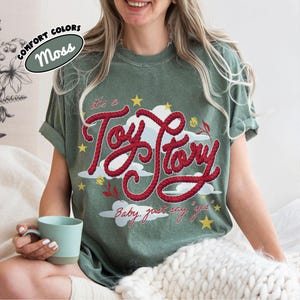 May include: A moss green t-shirt featuring a graphic design with the red rope-style text "it's a Toy Story" and "Baby, just say 'yes'" in red script. The design includes white clouds, yellow stars, and small red and yellow floral accents.