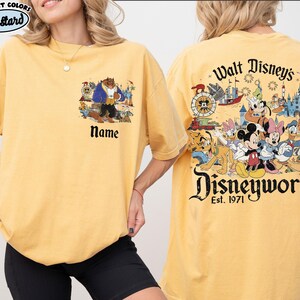 May include: Mustard-colored t-shirt with a Disney theme. The front features a Beauty and the Beast graphic with the word "Name" below. The back displays a Disney World graphic with various characters and the text "Walt Disney's Disneyworld Est. 1971."