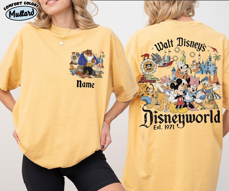 May include: Mustard-colored t-shirt with a Disney theme. The front features a Beauty and the Beast graphic with the word "Name" below. The back displays a Disney World graphic with various characters and the text "Walt Disney's Disneyworld Est. 1971."