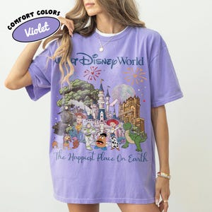 May include: Violet Comfort Colors t-shirt featuring a colorful graphic of Disney World characters and landmarks. The text "Walt Disney World" and "The Happiest Place On Earth" are also visible on the shirt.