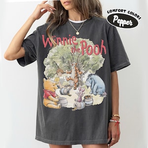 May include: Dark gray t-shirt featuring a graphic of Winnie the Pooh and friends under a tree. The design includes Winnie the Pooh, Tigger, Piglet, Eeyore, and Christopher Robin. The text "Winnie the Pooh" is in red.