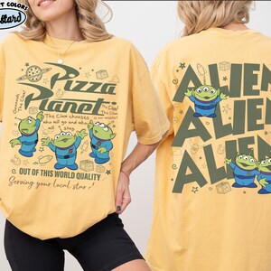 May include: Mustard-colored t-shirt featuring the Pizza Planet logo and three green aliens in blue suits. The front of the shirt has text that reads "Out of this world quality, serving your local star!" The back of the shirt has the word "ALIEN" repeated three times with alien graphics.