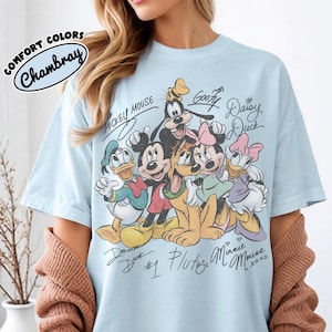May include: Light blue t-shirt featuring a graphic of Disney characters including Mickey Mouse, Minnie Mouse, Donald Duck, and Goofy. The design includes character signatures and the text "COMFORT COLORS Chambray".