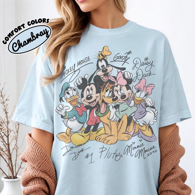 May include: Light blue t-shirt featuring a graphic of Disney characters including Mickey Mouse, Minnie Mouse, Donald Duck, and Goofy. The design includes character signatures and the text "COMFORT COLORS Chambray".