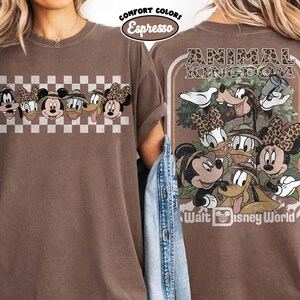 K&ouml;nnte beinhalten: Espressofarbenes T-Shirt mit einer Grafik von Disney-Charakteren. Die Vorderseite zeigt Gesichter von Charakteren auf einem Schachbrettmuster. Die R&uuml;ckseite hat ein Animal Kingdom-Design mit Charakteren und dem Text "Walt Disney World".