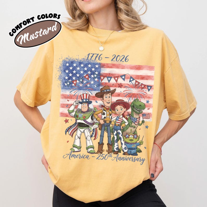 Może przedstawiać: Musztardowa koszulka z grafiką amerykańskiej flagi i postaciami z Toy Story. Projekt zawiera tekst "1776-2026" i "America - 250th Anniversary". Koszulka marki Comfort Colors.