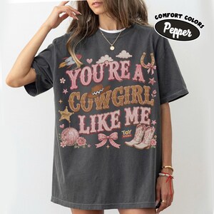Puede incluir: Una camiseta de manga corta gris carb&oacute;n con el texto "YOU'RE A COWGIRL LIKE ME" en rosa y marr&oacute;n. El gr&aacute;fico incluye un cohete, nubes, un sombrero de vaquero, una estrella de sheriff, una bola de discoteca, un lazo y botas con "TOY STORY".