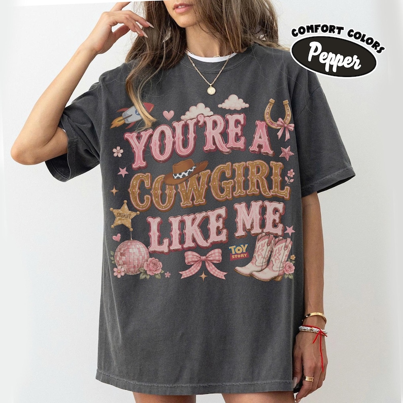 Puede incluir: Una camiseta de manga corta gris carb&oacute;n con el texto "YOU'RE A COWGIRL LIKE ME" en rosa y marr&oacute;n. El gr&aacute;fico incluye un cohete, nubes, un sombrero de vaquero, una estrella de sheriff, una bola de discoteca, un lazo y botas con "TOY STORY".
