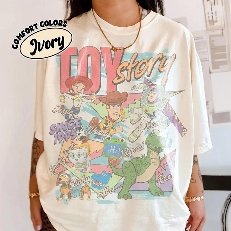 May include: An ivory t-shirt featuring a graphic print of Toy Story characters including Jessie, Woody, Buzz Lightyear, Rex, Slinky, and others. The design includes the text "TOY story", "SINCE 1995", and character names. A "COMFORT COLORS Ivory" label is visible.