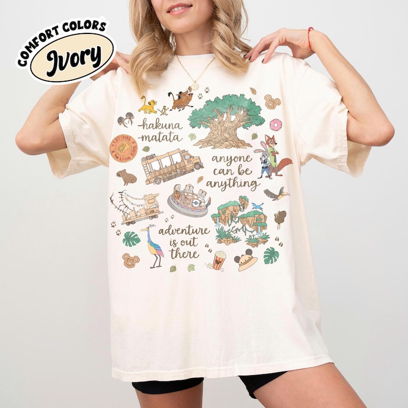 Puede incluir: Camiseta de color marfil con un dise&ntilde;o gr&aacute;fico colorido que presenta personajes de dibujos animados, un &aacute;rbol y las frases "hakuna matata" y "adventure is out there". La camiseta tambi&eacute;n tiene el texto "anyone can be anything".