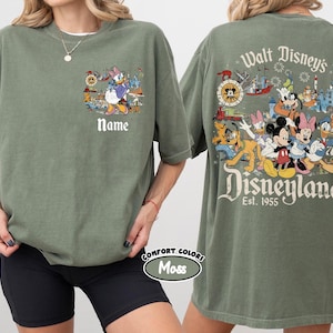 Peut inclure: T-shirt vert olive &agrave; th&egrave;me Disney. Le devant pr&eacute;sente Daisy Duck et Donald Duck avec le mot &laquo;&nbsp;Name&nbsp;&raquo;. Le dos affiche des personnages Disney et le texte &laquo;&nbsp;Walt Disney's Disneyland Est. 1955&nbsp;&raquo;.