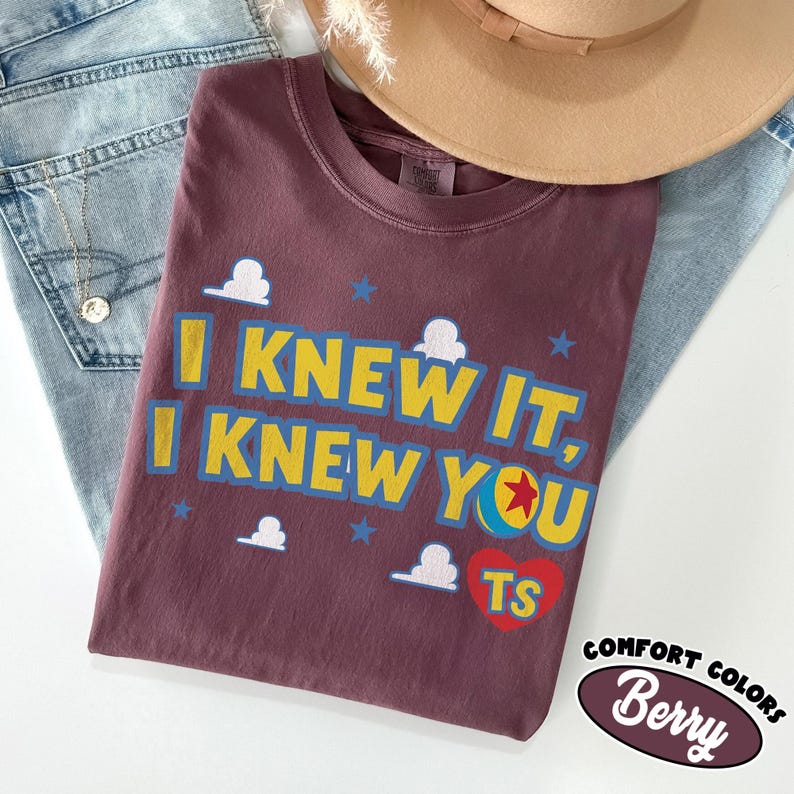 May include: A berry-colored t-shirt featuring the yellow text I KNEW IT, I KNEW YOU with a red heart containing the letters TS and a colorful ball graphic. The design includes small white cloud and blue star icons printed on the fabric.