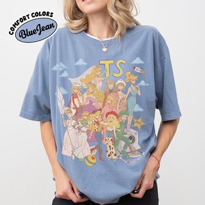 May include: A blue short-sleeve t-shirt featuring a graphic print of multiple animated characters in various outfits, including a space suit, cowgirl attire, and dresses. The text TS is printed in yellow above the characters, with a small rocket ship.