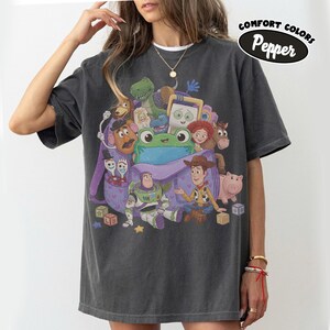 Pode incluir: Uma t-shirt cinza-carv&atilde;o com uma estampa gr&aacute;fica de v&aacute;rios personagens de brinquedos animados saindo de uma mochila roxa. O design inclui um sapo verde, um patrulheiro espacial, um cowboy e outras figuras. O texto Comfort Colors Pepper &eacute; vis&iacute;vel.