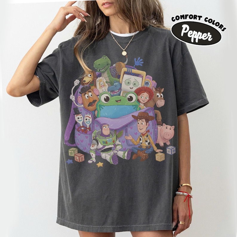Pode incluir: Uma t-shirt cinza-carv&atilde;o com uma estampa gr&aacute;fica de v&aacute;rios personagens de brinquedos animados saindo de uma mochila roxa. O design inclui um sapo verde, um patrulheiro espacial, um cowboy e outras figuras. O texto Comfort Colors Pepper &eacute; vis&iacute;vel.