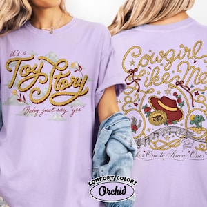 以下が含まれることがあります： ライトパープルのTシャツ。前面にはロープ風の文字で「it's a Toy Story Baby, just say "yes"」、背面には「Cowgirl like Me」の文字、カウボーイハット、音符、星が描かれたグラフィックがプリントされています。