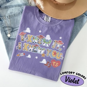 May include: A violet short-sleeved t-shirt featuring a graphic print with the text I KNEW IT, I KNEW YOU. The design includes cartoon elements like cowboy hats, boots, a rocket, stars, clouds, and a heart with the letters TS inside the print.