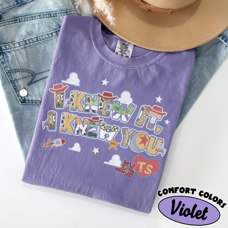 May include: A violet short-sleeved t-shirt featuring a graphic print with the text I KNEW IT, I KNEW YOU. The design includes cartoon elements like cowboy hats, boots, a rocket, stars, clouds, and a heart with the letters TS inside the print.