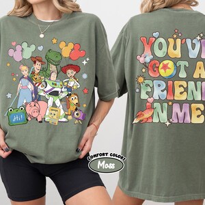 May include: A moss green t-shirt featuring a front graphic of Toy Story characters including Woody, Buzz Lightyear, Jessie, Rex, and Hamm. The back graphic displays the text "YOU'VE GOT A FRIEND IN ME" in colourful, bubble-style lettering.