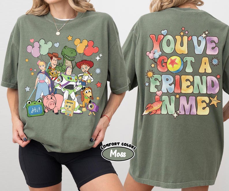 May include: A moss green t-shirt featuring a front graphic of Toy Story characters including Woody, Buzz Lightyear, Jessie, Rex, and Hamm. The back graphic displays the text "YOU'VE GOT A FRIEND IN ME" in colourful, bubble-style lettering.