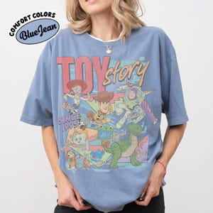 May include: A blue short-sleeve t-shirt featuring a graphic print of Toy Story characters including Woody, Buzz Lightyear, Jessie, Rex, and Slinky Dog. The design includes the text Toy Story, Since 1995, and character names. A Comfort Colors Blue Jean logo is visible.
