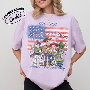 Może przedstawiać: Jasnofioletowa koszulka z grafiką przedstawiającą postacie z filmu Toy Story na tle amerykańskiej flagi. Koszulka zawiera napisy "1776-2026" i "America - 250th Anniversary". Koszulka jest oznaczona jako "Comfort Colors" i "Orchid".