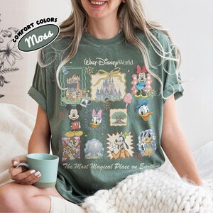 May include: Moss green t-shirt featuring a Walt Disney World design with various cartoon characters and park icons. The shirt includes the text "The Most Magical Place on Earth" and "Comfort Colors".