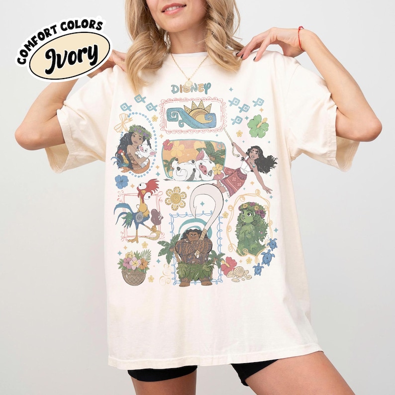 May include: Ivory-coloured t-shirt featuring Disney characters from the movie Moana. The shirt has a crew neck and short sleeves. The graphic includes illustrations of Moana, Maui, Hei Hei, Pua, and other characters. The text "Disney" is printed at the top.