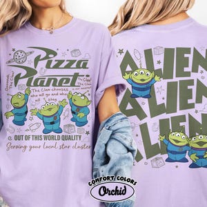 May include: A light purple t-shirt featuring the Pizza Planet logo and three green aliens in blue uniforms. The text reads "Out of this world quality" and "Serving your local star cluster."